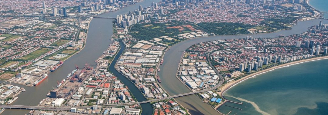 A wide, zoomed-out aerial photograph of Jakarta's coastline, showing the vast urban sprawl of the city meeting the Java Sea, with rivers and canals flowing into the ocean.