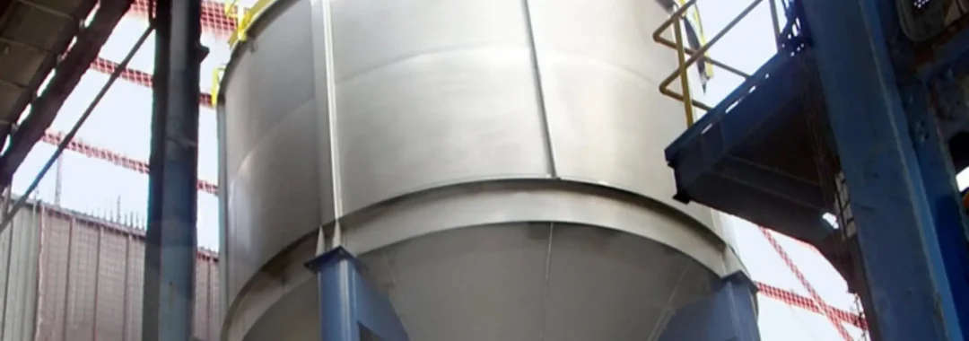 A low-angle shot of a large, cylindrical, stainless steel industrial decant tank with a conical bottom, supported by blue steel beams inside a factory setting. A yellow safety railing runs along the top edge of the tank.