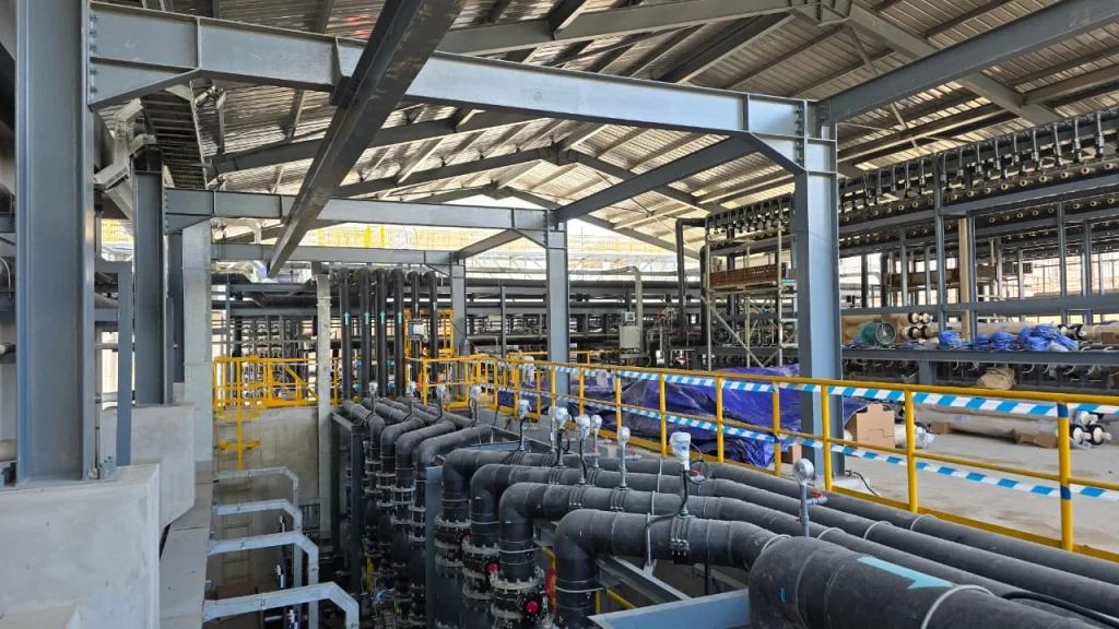 A wide interior shot of the PT GLS desalination plant, showcasing the complex network of piping in the foreground and the multiple racks of SWRO membrane vessels (trains) in the background.